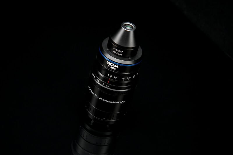 A black Laowa 5-10x Ultra Macro APO lens stands vertically on a reflective black surface.