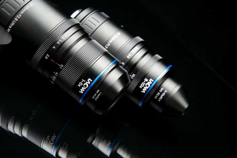 Two Laowa macro lenses are positioned diagonally on a black reflective surface, with their reflections visible below them.