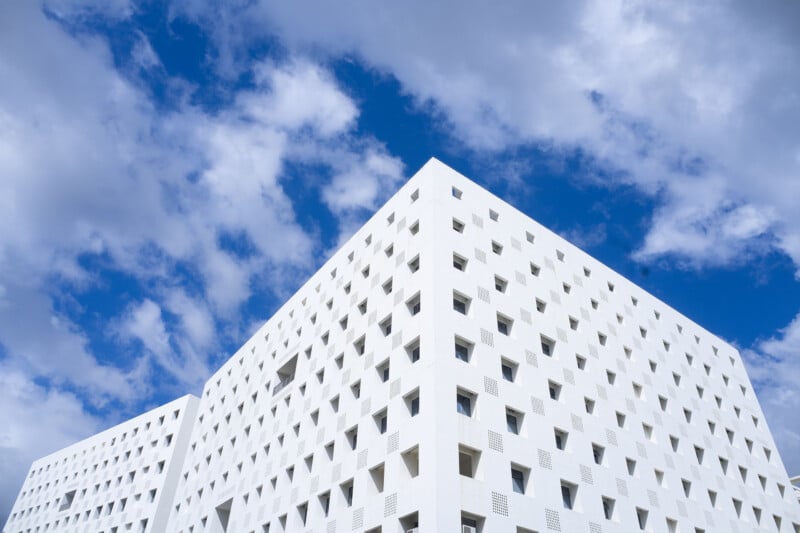 A modern white building with many small, square windows stands against a bright blue sky with scattered clouds. The building's geometric design and clean lines create a striking visual effect.