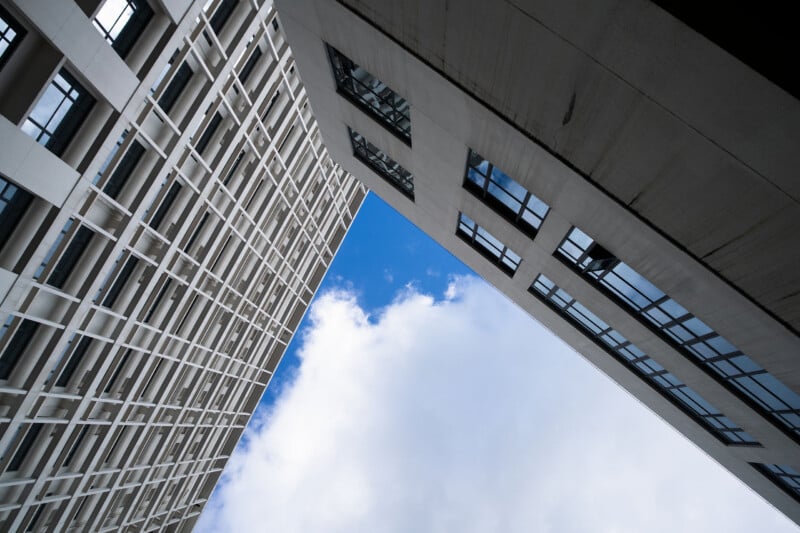 Two modern buildings form an angular frame, creating a sharp V-shape that opens up to blue sky and white clouds above. The photo is taken from a low angle, looking directly upward.