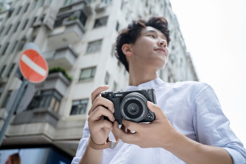 A person stands outdoors holding a Nikon camera, looking off to the side, with tall buildings and a no-entry sign visible in the background.