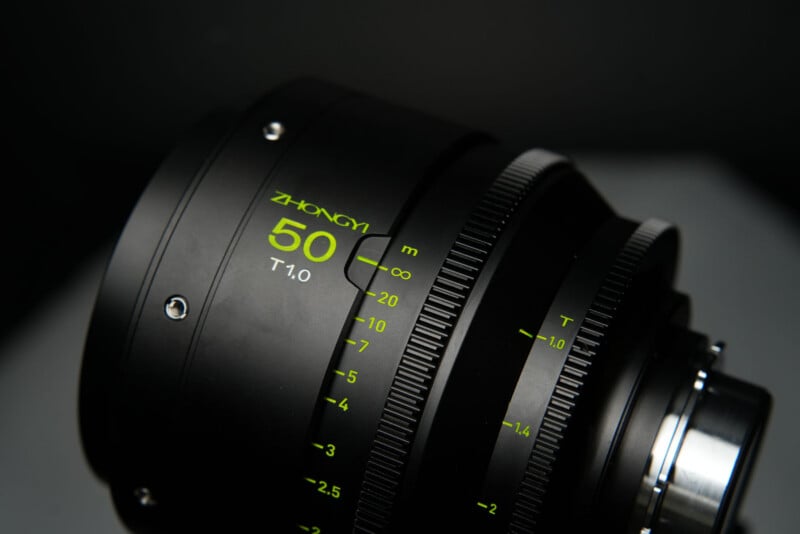 Close-up of a Zhong Yi camera lens with "50 T1.0" and distance markings in bright green text on a black metal body. The lens focus and aperture rings are also visible.
