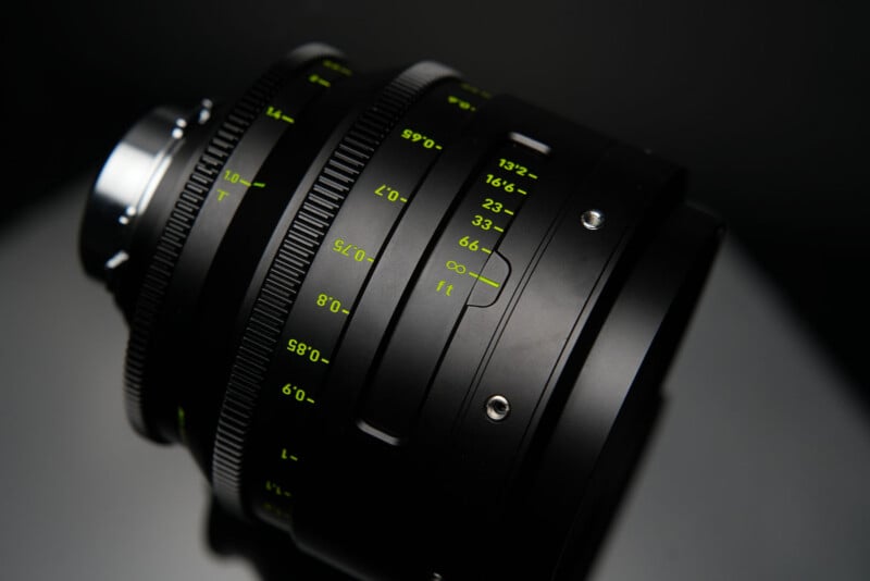 A close-up of a black camera lens with yellow-green markings for focus and aperture settings, placed on a reflective surface against a dark background.