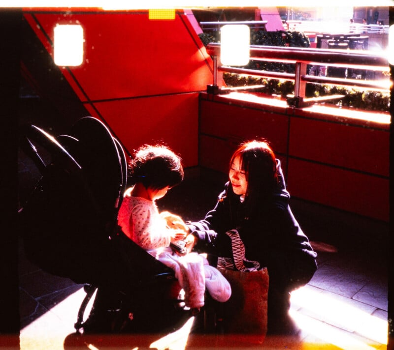 A woman kneels beside a child seated in a stroller, both illuminated by warm sunlight. The scene is set against a red wall with railings, and there are bright light flares in the image.