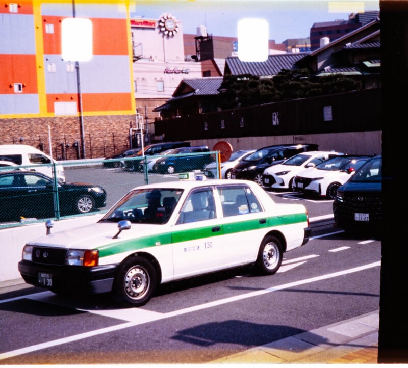 A white and green taxi is parked on a city street next to a row of parked cars, with colorful buildings and a clock visible in the background. The scene appears to be in Japan.