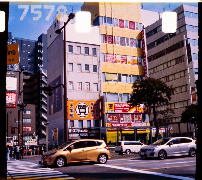 Busy urban street in Japan with cars crossing at a crosswalk, surrounded by tall buildings adorned with colorful signs and advertisements, including yellow and red storefronts. Trees and pedestrians are visible on the sidewalks.