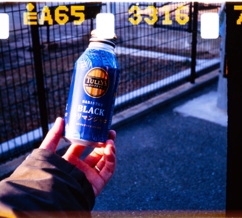 A hand holds a blue can of Tully’s Coffee Barista’s Black outdoors; the background shows a paved area and a metal fence, with film strip numbers visible at the top and black film borders framing the image.