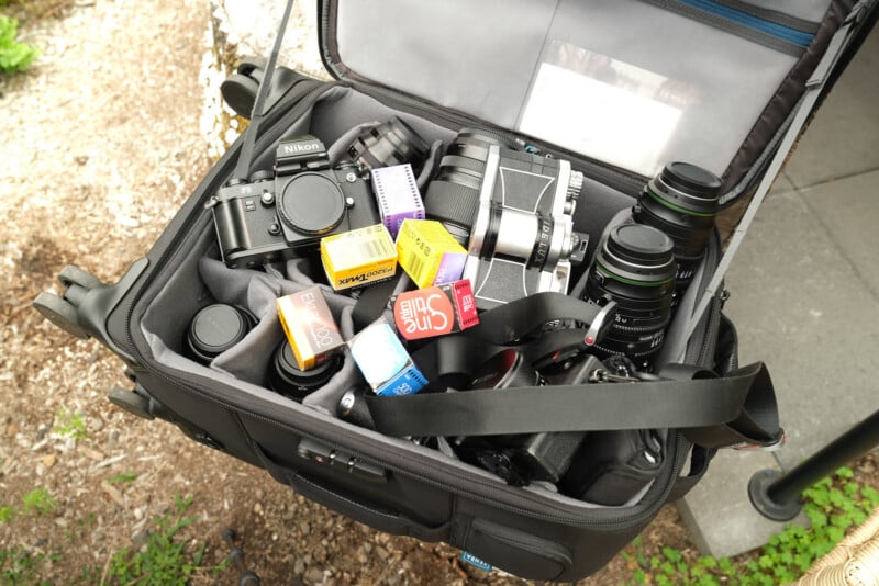 An open camera bag containing film cameras, camera lenses, and several boxes of 35mm film in various colors and brands. The bag is organized with padded sections for the equipment.