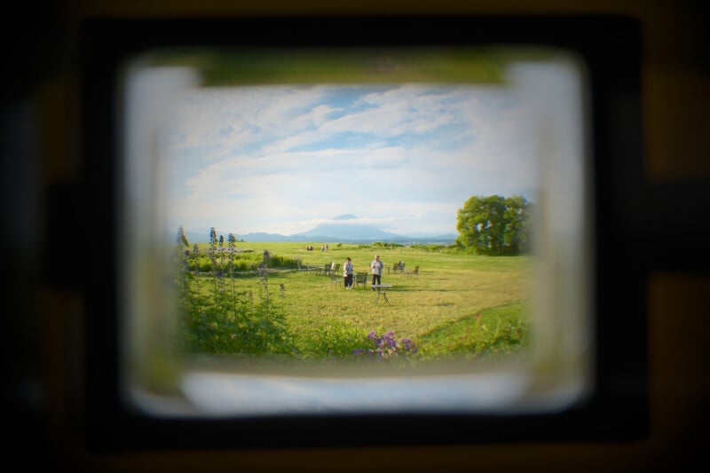 View of a grassy field with people and distant mountains, seen through a blurry rectangular frame, suggesting the perspective of looking through a window or camera viewfinder.