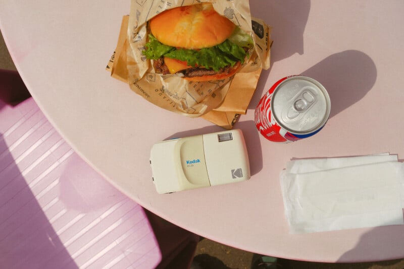 A wrapped burger, an open can of soda, a vintage Kodak camera, and napkins are on a round pink table with a pink chair beside it, viewed from above.