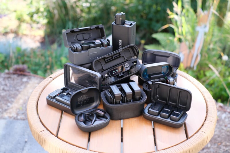 A round table outdoors displays various black electronic devices and cases, including headphones, chargers, and batteries, with green plants blurred in the background.