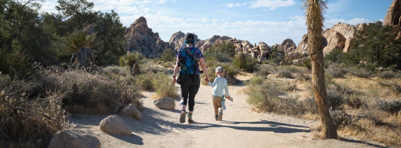 A woman and a child holding hands walk along a sandy trail through a desert landscape with rocks, bushes, and Joshua trees under a partly cloudy sky.