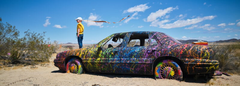 A person in a yellow shirt and white hat stands next to a car covered in colorful paint splatters, parked in a desert landscape under a bright blue sky with scattered clouds.