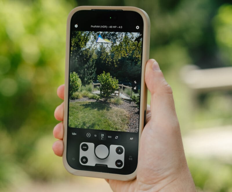 A hand holds a smartphone displaying a camera app, capturing a small bridge and greenery in an outdoor park setting with abundant sunlight.