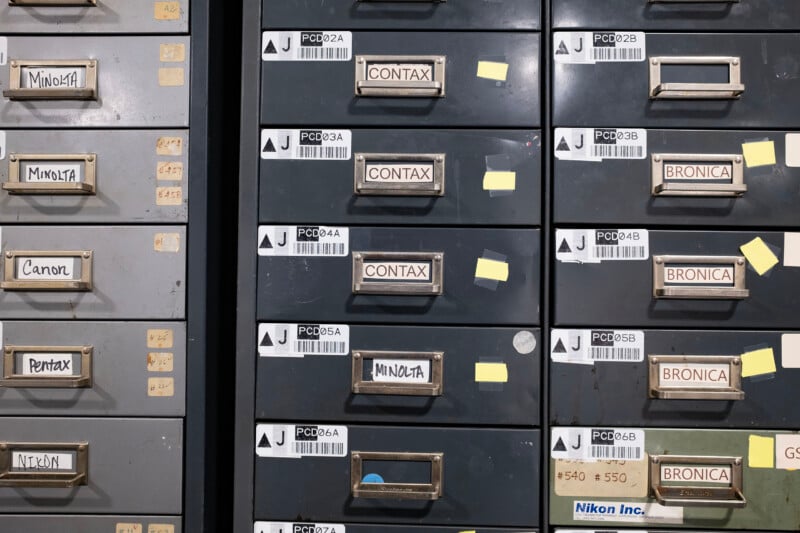 A metal filing cabinet with multiple drawers labeled with brand names including Minolta, Contax, Bronica, Canon, Pentax, and Nikon.
