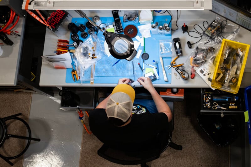 A person wearing a baseball cap sits at a workbench repairing electronic components surrounded by tools, lenses, and parts in a workshop.