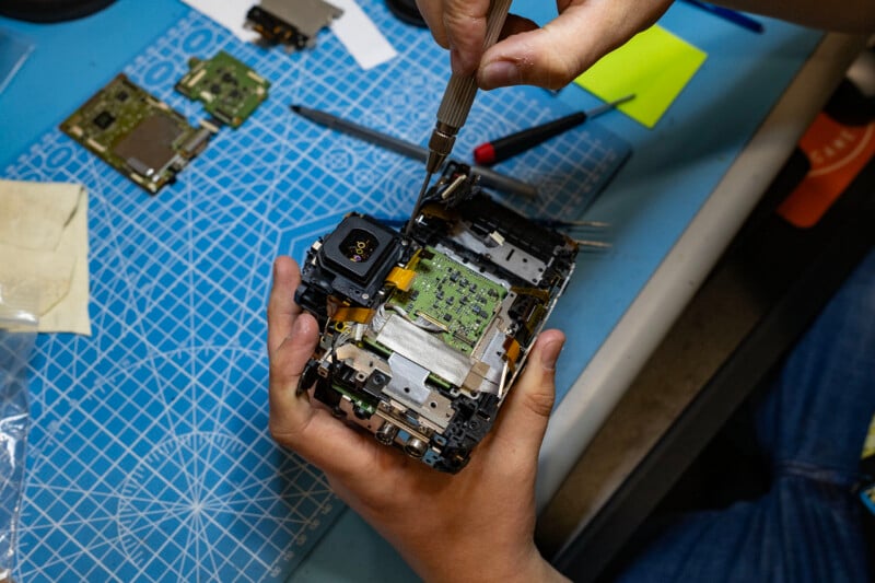 A person uses a precision screwdriver to work on the internal components of a disassembled camera held in their hands on a blue cutting mat.
