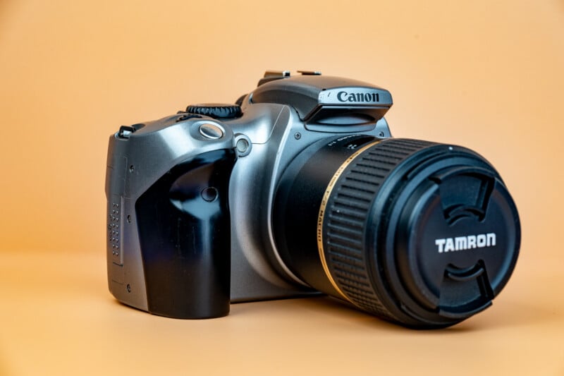 A Canon DSLR camera with a Tamron lens attached, placed on a smooth, light orange background. The camera is angled slightly to the left, prominently displaying the Canon and Tamron brand names.