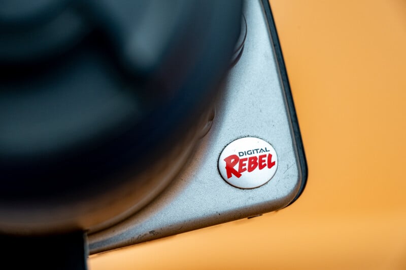 Close-up of a camera body corner with a round button labeled "DIGITAL REBEL" in red and black text, set against a blurred orange background. Part of a camera lens is visible on the left.