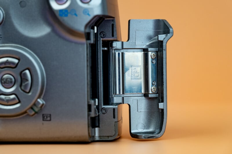 Close-up of a digital camera’s open memory card slot with a CompactFlash card partially inserted, set against an orange background. Camera buttons and part of the body are visible on the left.