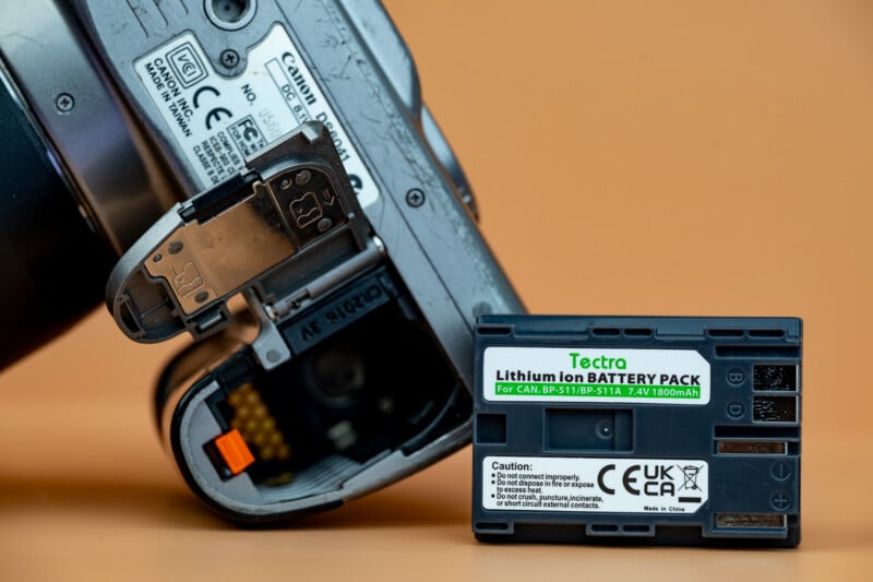 A close-up of a camera with its battery compartment open, next to a Tectra lithium ion battery pack labeled for Canon cameras, against an orange background.