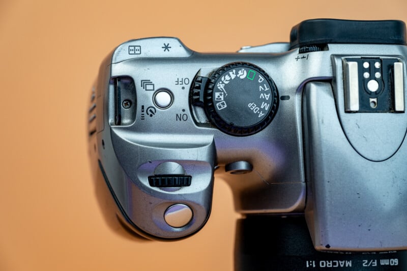 Top view of a silver digital camera showing its mode dial, power switch, and various control buttons against an orange background. The mode dial includes options like auto, manual, and scene settings.