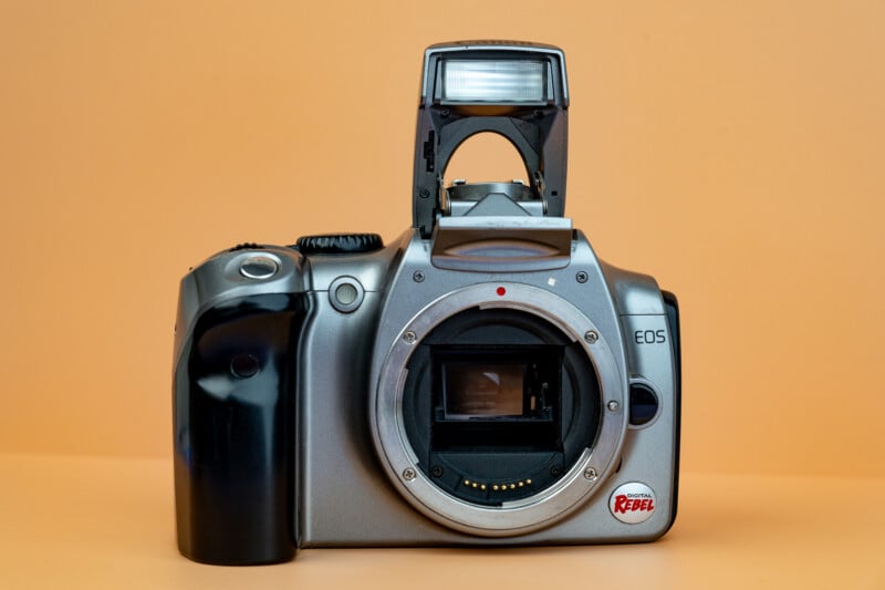 A silver Canon EOS Rebel digital camera without a lens, showing its open lens mount and raised pop-up flash, against a plain peach background.