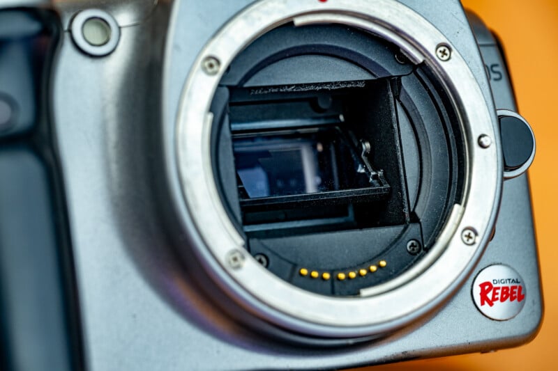 Close-up of a DSLR camera body without a lens, showing the exposed sensor and lens mount. The camera has a "Digital Rebel" logo on the lower right.
