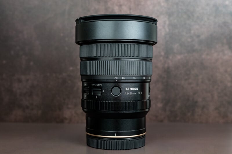 A Tamron 12-20mm F2.8 camera lens stands upright on a flat surface with a muted, textured background.