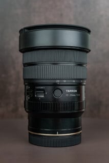 A black Tamron 12-20mm F2.8 camera lens stands upright on a flat surface with a brown gradient background, showing detailed focus and custom controls on its side.