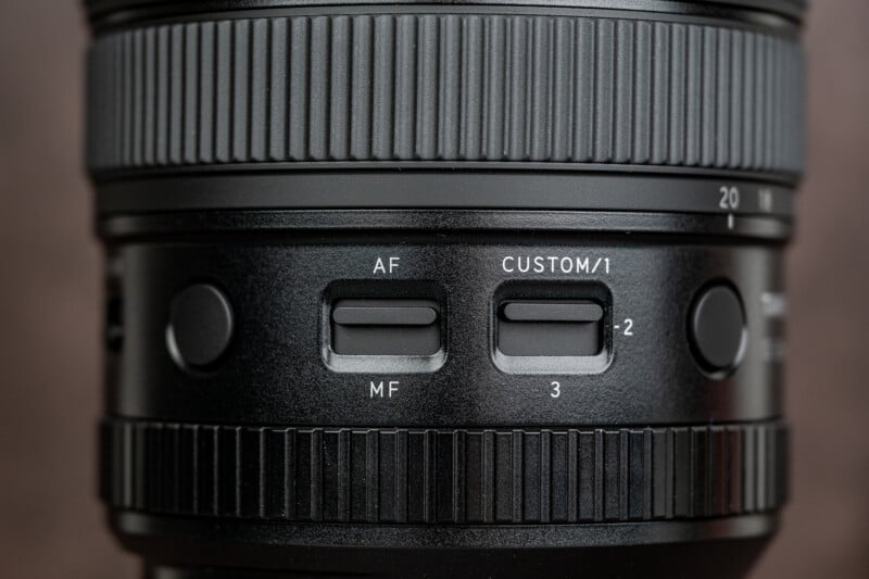 Close-up of a camera lens showing textured focus rings, two rectangular buttons labeled AF/MF and CUSTOM/1-2-3, and two round buttons on either side, with detailed markings and grooves visible.