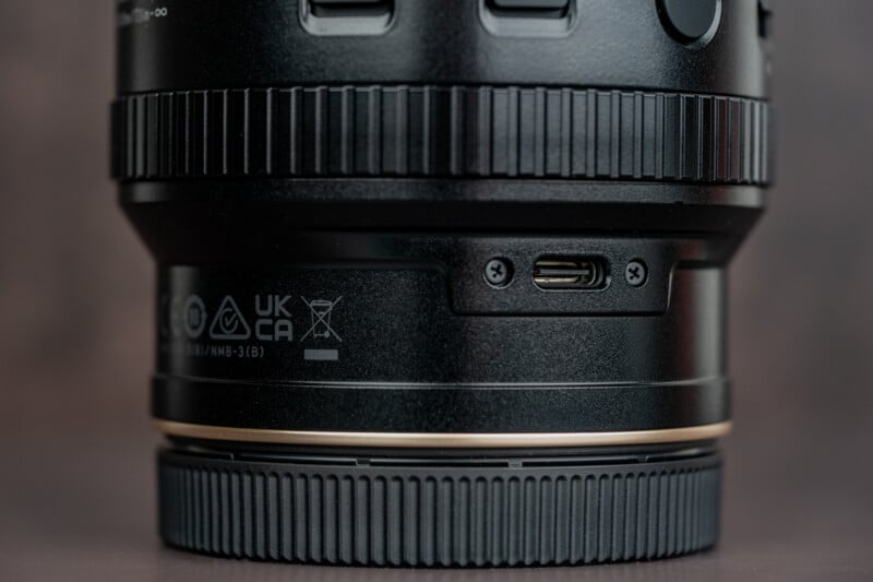 Close-up of a black camera lens showing a USB-C port, various certification symbols, textured grip rings, and part of the lens mount.