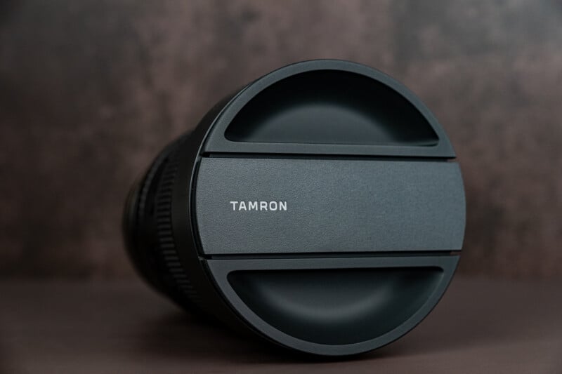 A close-up of a Tamron camera lens with its lens cap on, placed on a brown surface against a blurred brown background.