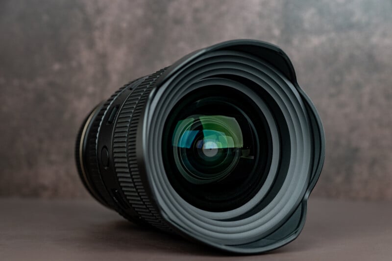 Close-up view of a black camera lens with a petal-shaped hood facing the viewer, set against a blurred brownish background.