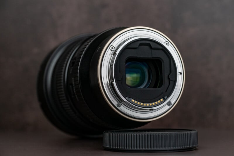 A close-up of a camera lens laying on its side with its rear lens cap off, showing the metal lens mount and electronic contacts against a dark, blurred background.