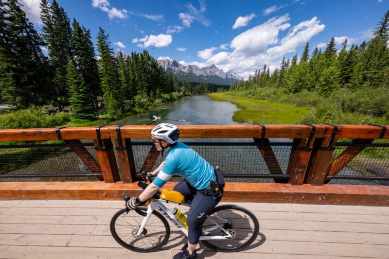 A person in blue cycling gear rides a bike across a wooden bridge over a river, surrounded by pine trees and mountains under a bright blue sky with white clouds.