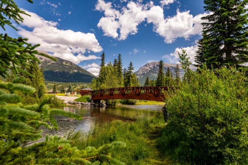 A wooden bridge crosses a clear creek surrounded by lush green trees, with tall mountains and a partly cloudy blue sky in the background.