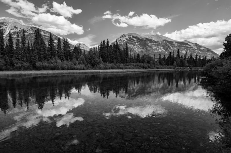 Calm lake reflecting trees, mountains, and clouds under a partly cloudy sky, with pine trees along the shoreline and mountains rising in the background. The image is in black and white.