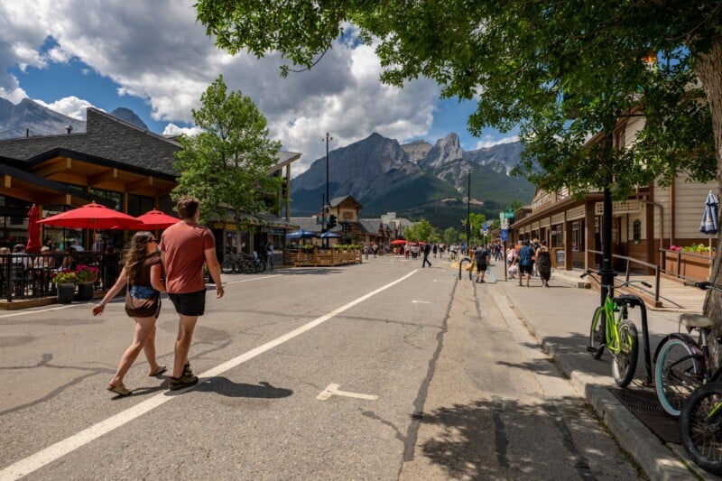 A couple walks hand in hand across a sunny street lined with shops and restaurants, with mountains and clouds in the background. People stroll, and bicycles are parked nearby.