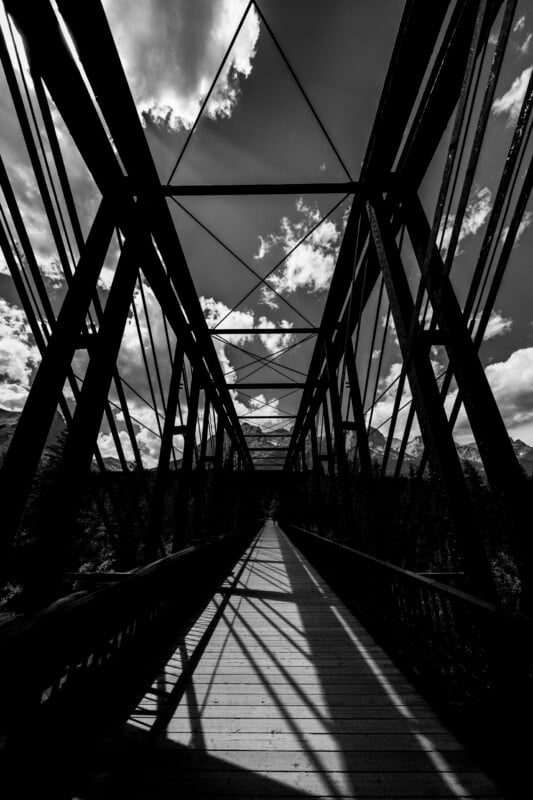 Black-and-white photo of a metal bridge, viewed from the entrance. The sun casts dramatic shadows of the bridge’s structure on the wooden walkway, and clouds drift in the sky above.