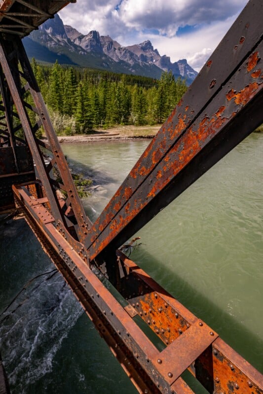 Rusty steel bridge beams over a green river, with a forest and tall, rocky mountain peaks under a partly cloudy sky in the background.