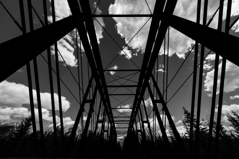 Black and white photo looking up at a metal truss bridge with clouds and sky visible through the structure, and trees lining the sides at the bottom.