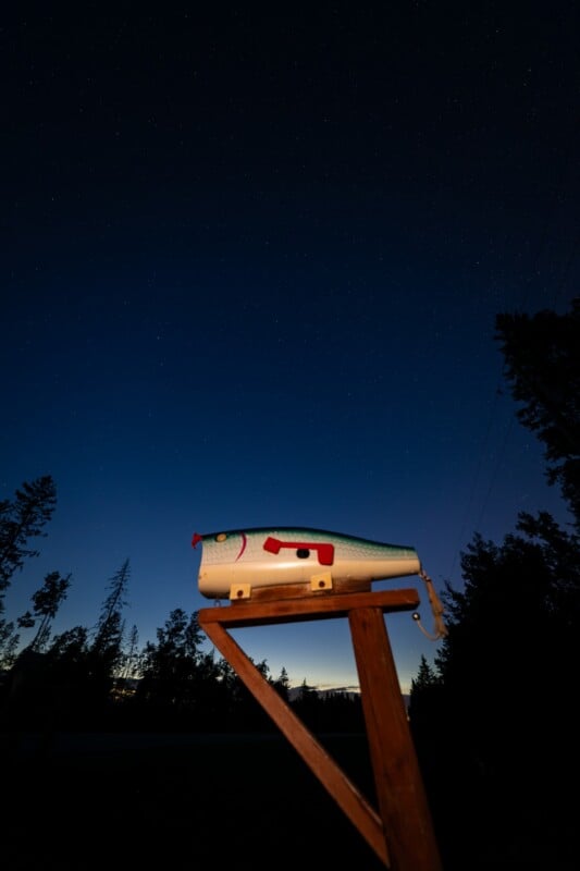 A white mailbox with red accents stands on a wooden post against a dark, starry night sky. Silhouetted trees frame the scene, with a faint glow at the horizon.