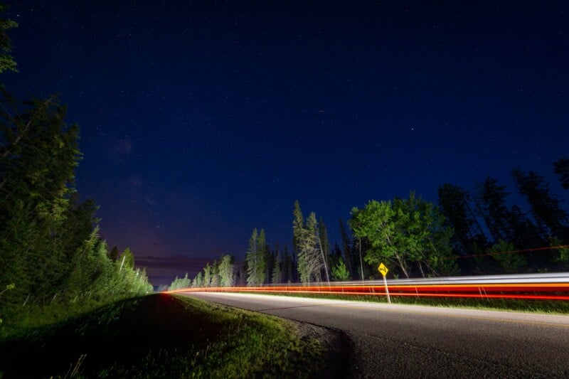 A curving road at night with trees on both sides, starry sky above, and red and white light trails from passing vehicles.
