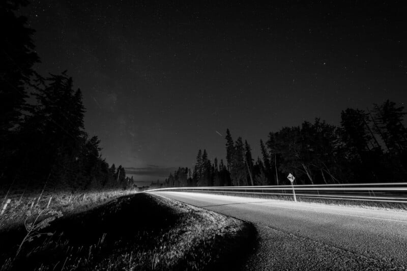 A black-and-white photo of a curving road at night, with light trails from passing cars. Dense trees line both sides of the road, and stars are visible in the sky above.