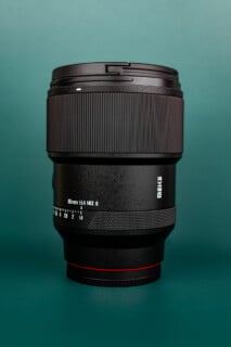 A black Meike 85mm f/1.4 camera lens stands upright on a green surface with a green background.