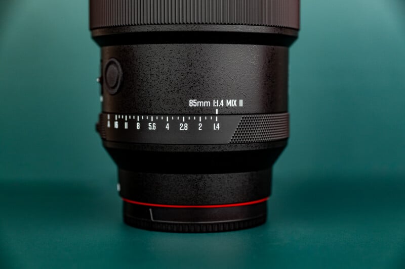 A close-up of a camera lens against a teal background, showing the label "85mm f/1.4 MIX II" and a focus distance scale.