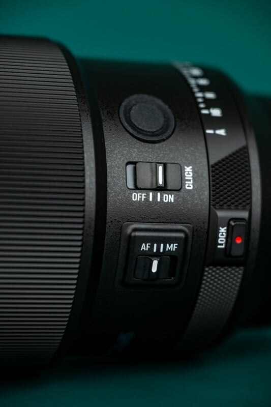 Close-up of a camera lens showing focus switch (AF/MF), a power switch (OFF/ON), a dial, and a “LOCK” slider with a red indicator. The background is teal.