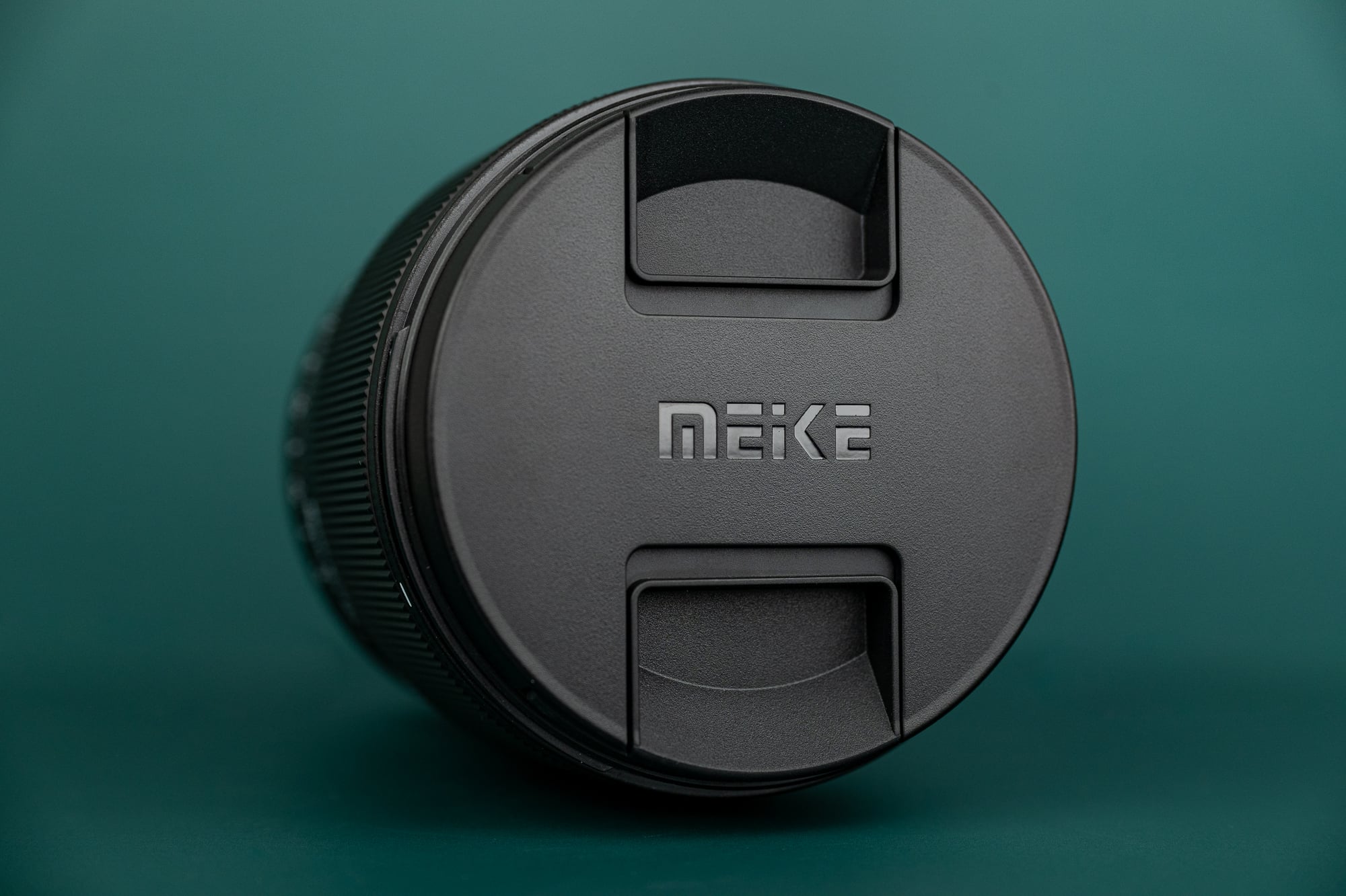 A close-up of a Meike camera lens with its lens cap on, placed against a green background.