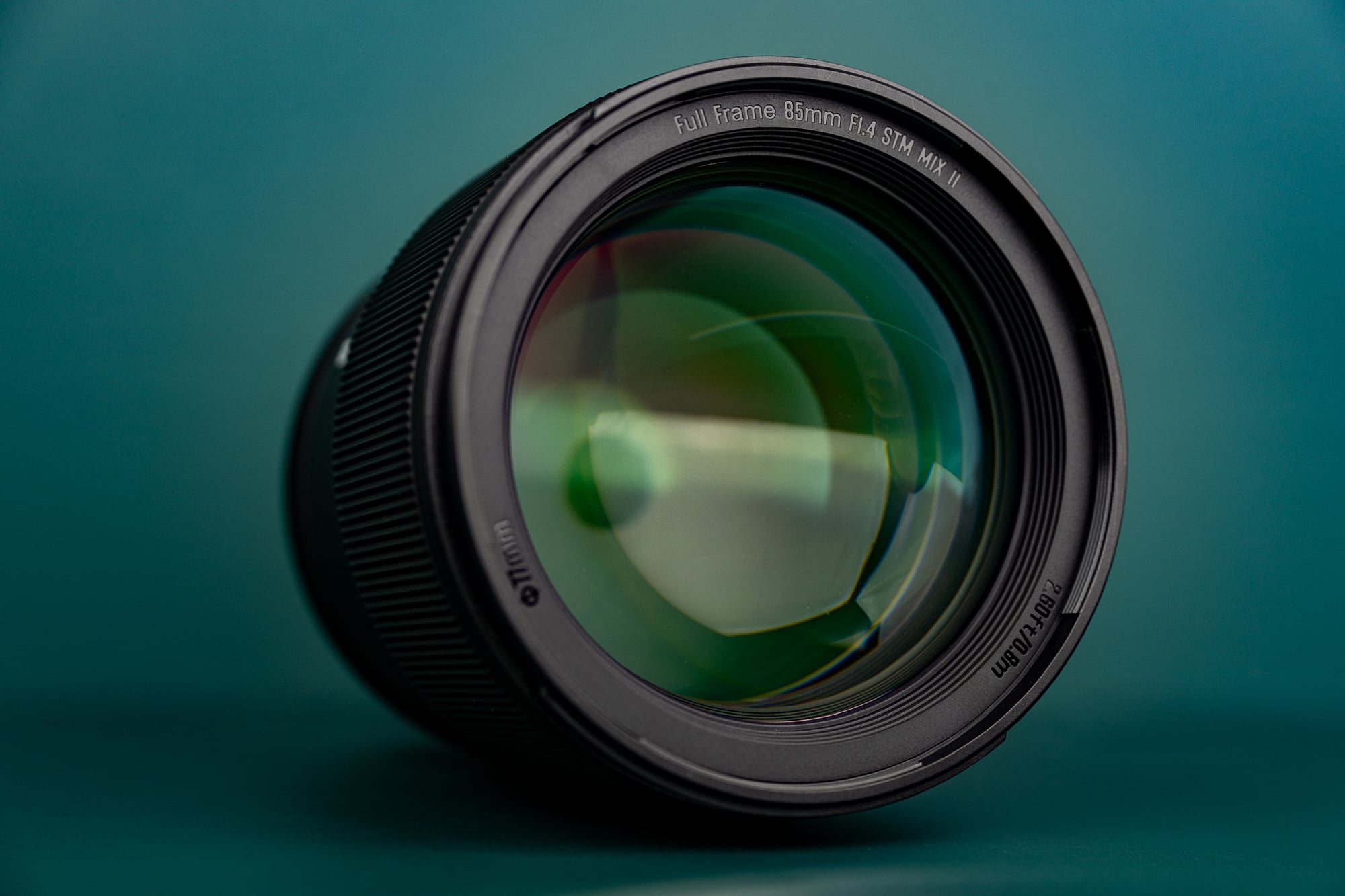 A close-up of a camera lens with visible reflections on the glass, set against a teal background. The lens markings indicate it is a Full Frame 85mm f/1.4 STM lens.